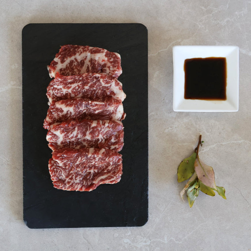 Raw beef yakiniku slices on a black board with a small bowl of soy sauce on a light gray surface.