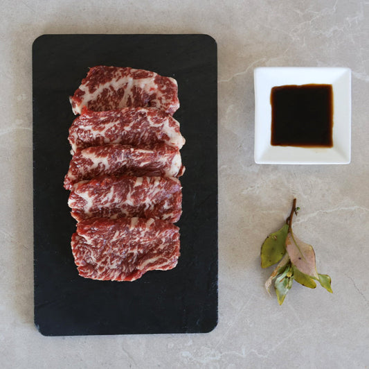 Raw beef yakiniku slices on a black board with a small bowl of soy sauce on a light gray surface.
