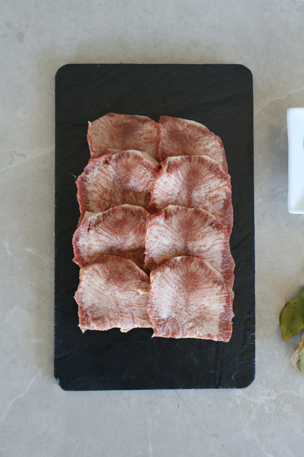 Sliced raw wagyu tongue on a black slate board on a light gray surface