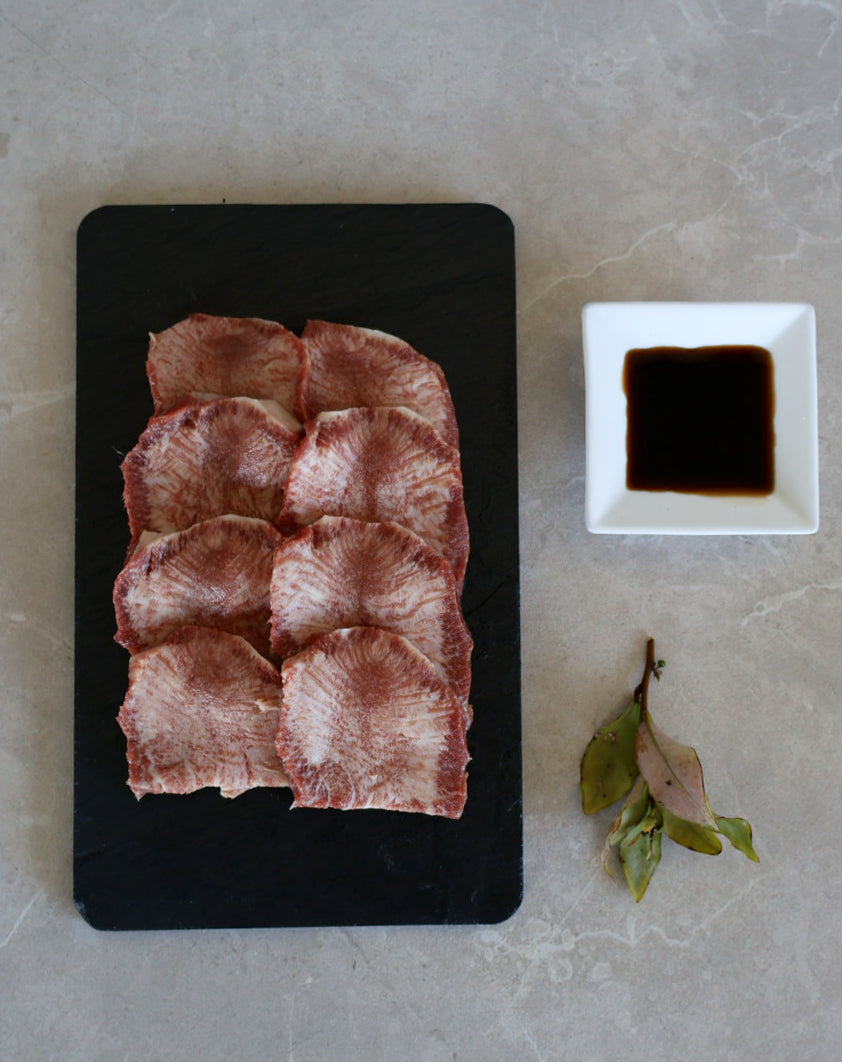 Raw wagyu tongue a black board with a small bowl of liquid and a leaf on a gray surface