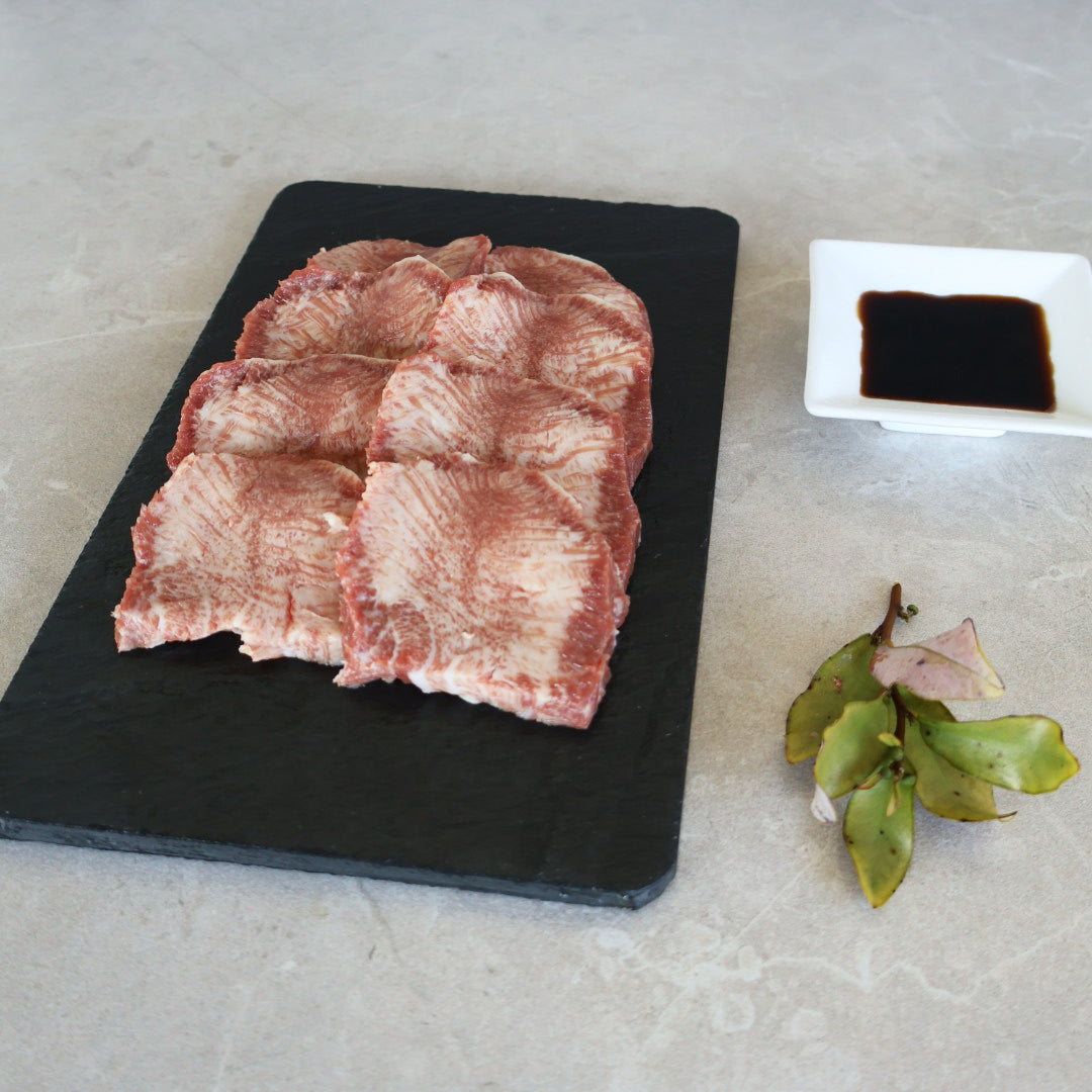 Raw wagyu beef tongue on a black cutting board with a small container of sauce and leaves on a light gray surface.