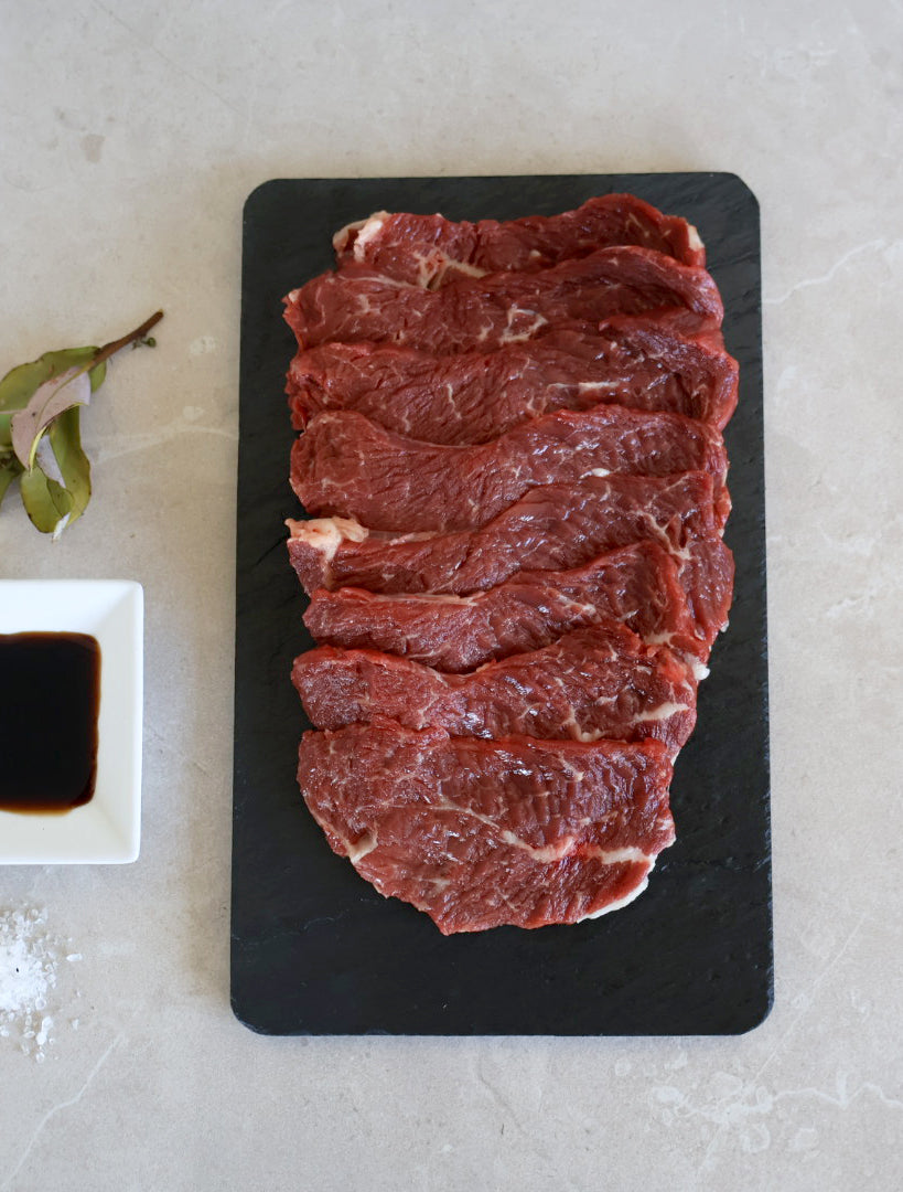 Raw Wagyu Beef Rump Yakiniku slices on a black slate board with a small bowl of sauce on a light gray background