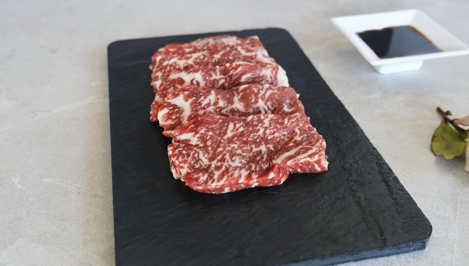 Raw marbled sirloin wagyu Yakiniku on a black slate board with a small bowl of sauce in the background.